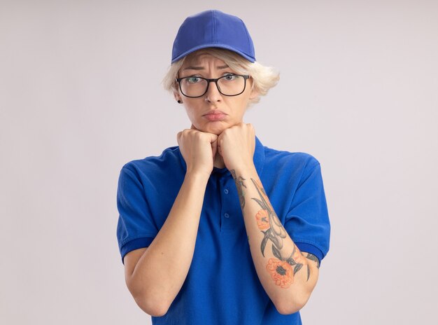 Расстроенная молодая женщина-доставщик в синей форме и кепке с грустным выражением лица смотрит на белую стену
