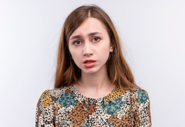 Upset young girl with long hair wearing colorful dress looking at camera with unhappy face 