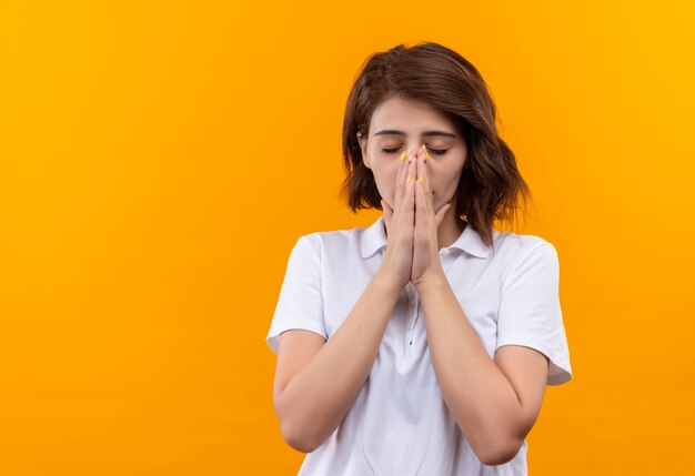 Upset young girl with short hair wearing white polo shirt holding hands witj closed eyes with hope expression