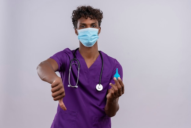 Расстроенный молодой красивый темнокожий доктор с кудрявыми волосами в фиолетовой униформе со стетоскопической маской для лица показывает большие пальцы вниз и держит клизму