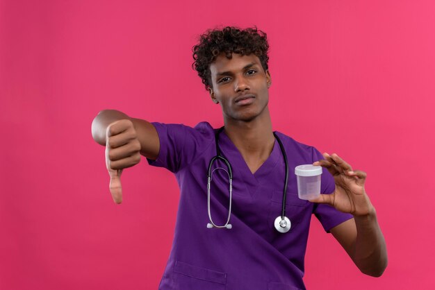 An upset young handsome dark-skinned doctor with curly hair wearing violet uniform with stethoscope showing thumbs down while holding plastic specimen jar 