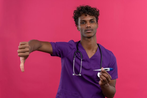Расстроенный молодой красивый темнокожий доктор с кудрявыми волосами в фиолетовой форме со стетоскопом показывает большие пальцы руки вниз и держит пластиковый термометр