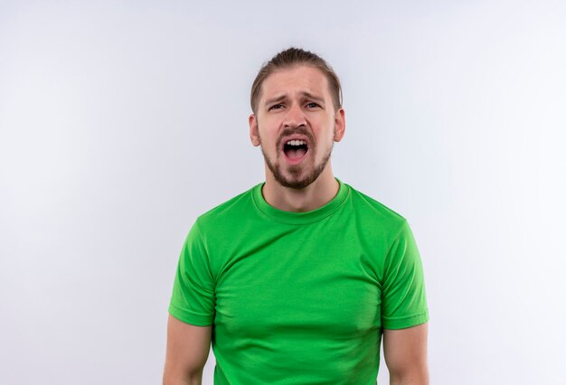 Upset young handsome man in green t-shirt shouting standing over white background