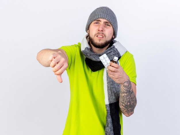 Расстроенный молодой больной человек в зимней шапке и шарфе показывает палец вниз и держит лекарство в стеклянной бутылке и упаковке медицинских таблеток, изолированных на белой стене