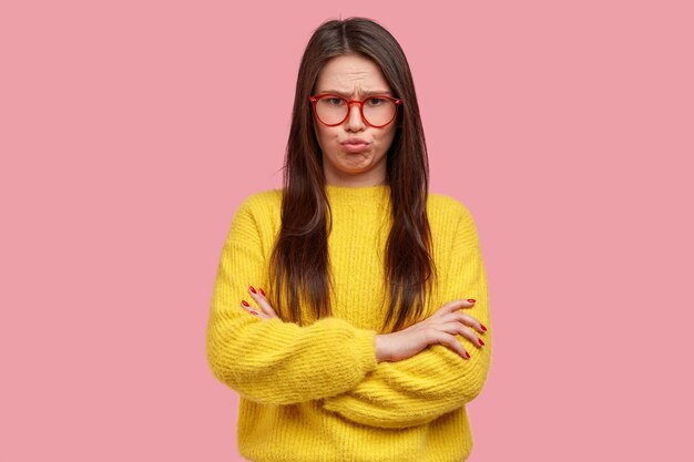 Upset young lady pouts lips from sadness, has dissatisfied expression, keeps arms folded, feels offended