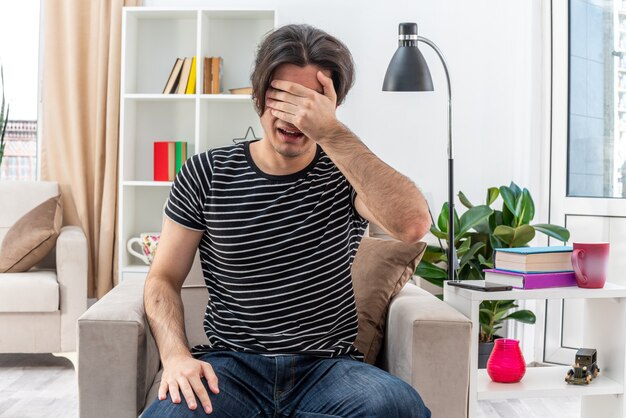 Upset young man in casual clothes covering eyes with hand sitting on the chair in light living room