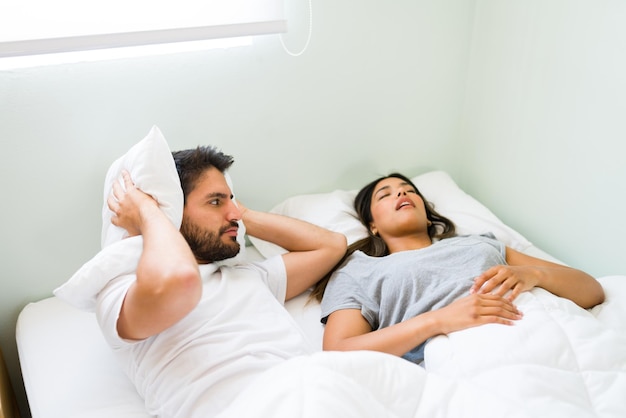 Upset young man covering his ears with a pillow and feeling angry with his snoring girlfriend while lying in bed
