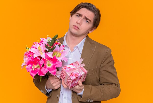 Расстроенный молодой человек с подарком и букетом цветов смотрит вперед с грустным выражением лица и собирается поздравить с международным женским днем, стоя над оранжевой стеной