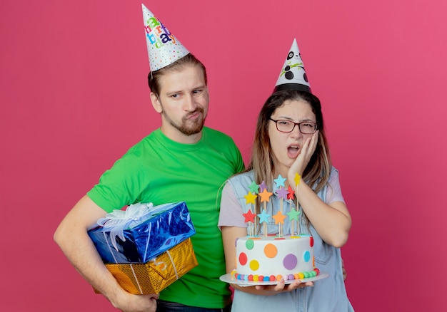 Бесплатное фото Расстроенный молодой человек в праздничной шляпе держит подарочные коробки, стоя с недовольной молодой девушкой в праздничной шляпе, кладет руку на лицо, держа именинный торт, изолированный на розовой стене