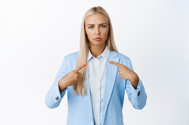 Upset young office lady pointing fingers at herself frowning and pouting displeased having doubts standing in blue suit over white background