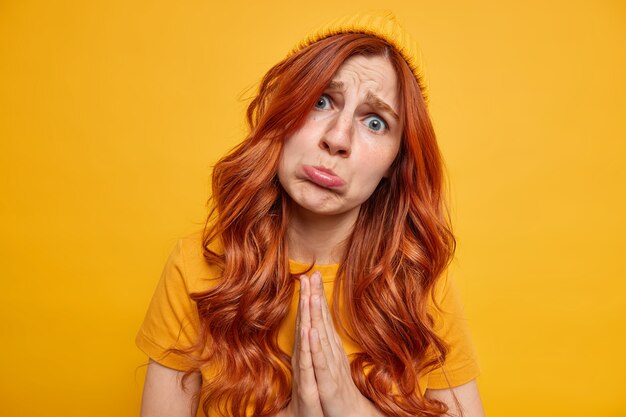 Upset young redhead woman purses lips keeps palms in pray begs you about something with pitiful expression looks hopeful dressed in casual yellow clothes has needy hopeless face stands indoor