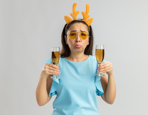 Расстроенная молодая женщина в синем топе в забавной оправе с оленьими рогами и в желтых очках с двумя бокалами шампанского смотрит с грустным выражением лица, поджав губы