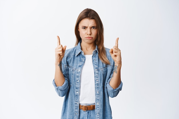 Upset young woman complain on unfair situation, frowning and sulking angry, pointing fingers up at disappointing offer, standing against white wall