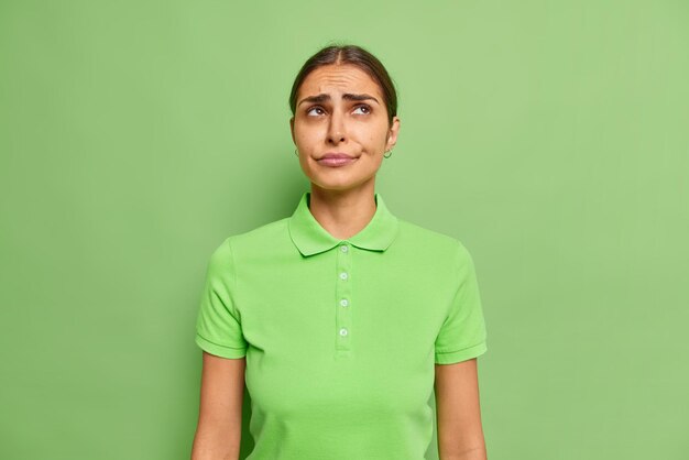 Upset young woman in despair regrets something purses lips concentrated above frowns face dressed in casual t shirt isolated over green wall