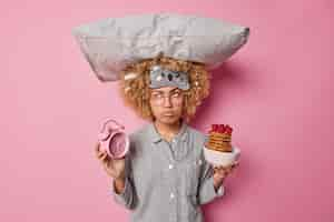 Free photo upset young woman dressed in pajama carries pillow on head holds alarm clock and bowl of pancakes with raspberries has sad pensive expression isolated over pink background morning time concept