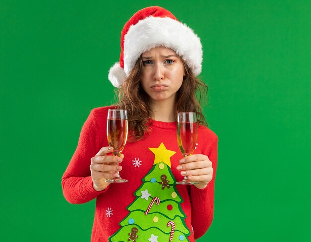 Расстроенная молодая женщина в красном рождественском свитере и шляпе санта-клауса, держащая два бокала шампанского, смотрит в камеру с грустным выражением лица, поджимая губы, стоя на зеленом фоне