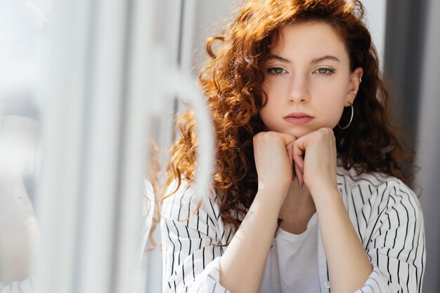 Upset young woman in shirt sitting on windowsill at home