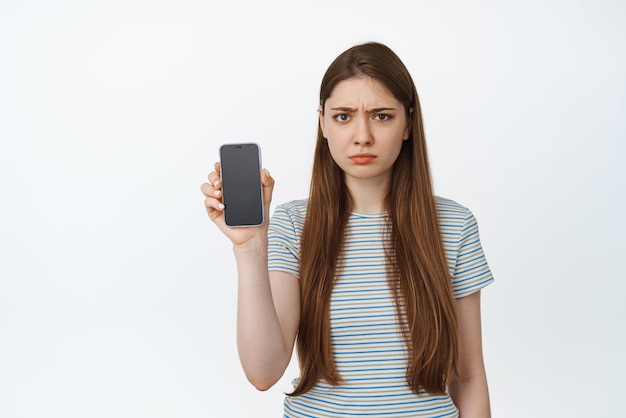 彼女の電話の画面を表示し、眉をひそめている若い女性がスマートフォンを保持している怒っている若い女性は、白い背景に対して悲しい怒っている顔が立っているモバイル画面を示しています