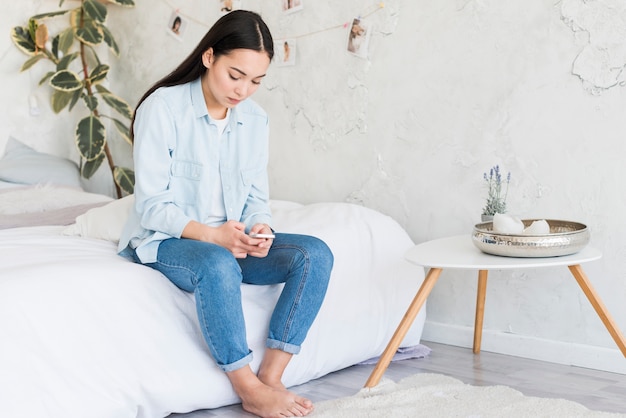 Upset young woman sitting on bed with mobile phone