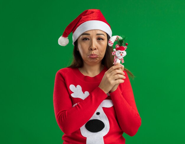 Upset young woman wearing christmas santa hat and red sweater holding christmas candy cane looking at camera with sad expression pursing lips  standing over green background