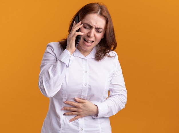 Upset young woman in white shirt talking on mobile phone crying standing over orange wall