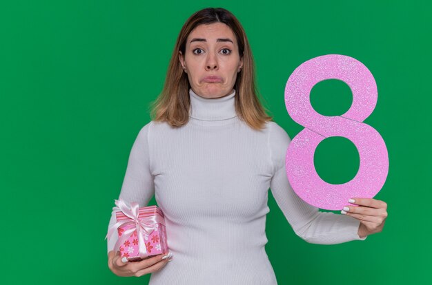 upset young woman in white turtleneck holding number eight and present