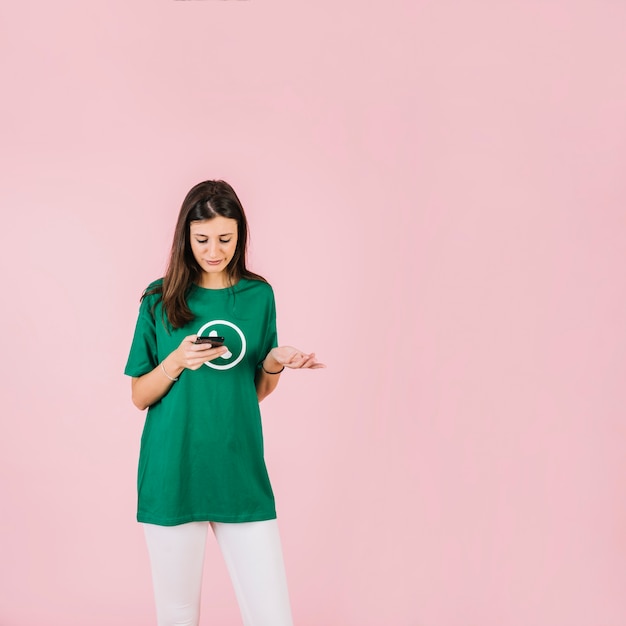 Upset young woman with smartphone shrugging against pink background