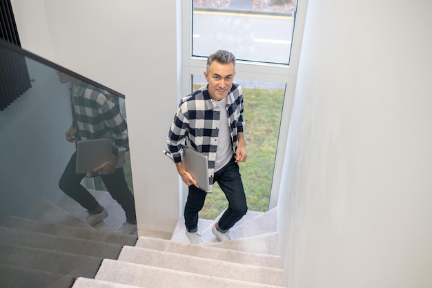 Foto gratuita al piano superiore vista dall'alto di un uomo di mezza età sorridente con un laptop che sale scale leggere