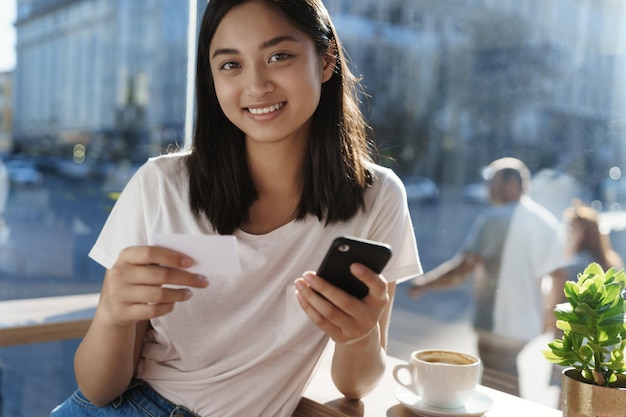 Urban lifestyle city people concept Lovely happy smiling asian girl in casual white tshirt hold smartphone and credit card sit near cafe windows at coffee table behind passersby walking street