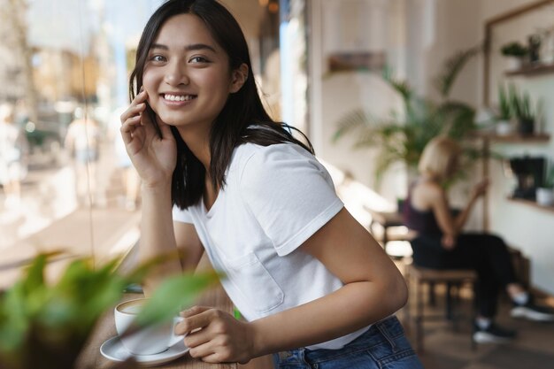 Urban lifestyle millennial and hipster concept Attractive lovely and cute asian girl student looking sideways as sitting in cafe near window talking to friend as drink coffee and enjoy dayoff