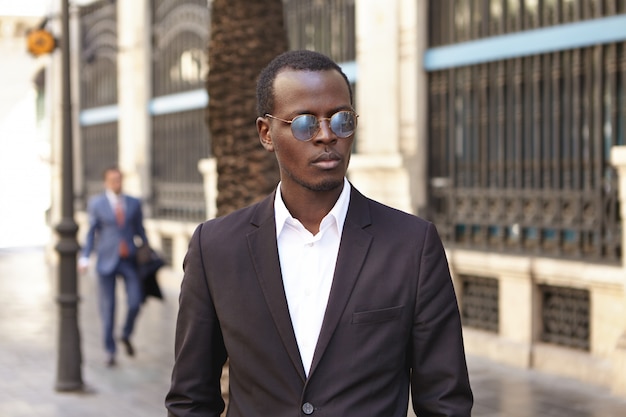 Urban outdoor portrait of confident serious young dark-skinned entrepreneur wearing stylish round shades and formal suit standing on street against office building