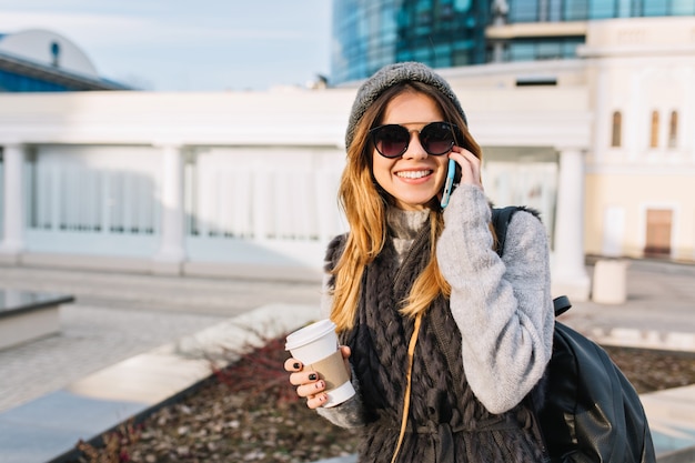 Urban stylish portrait of amazing joyful young woman in warm woollen sweater, knitted hat, modern sunglasses walking in sunny city centre with coffee to go. Cheerful emotions, place for text.