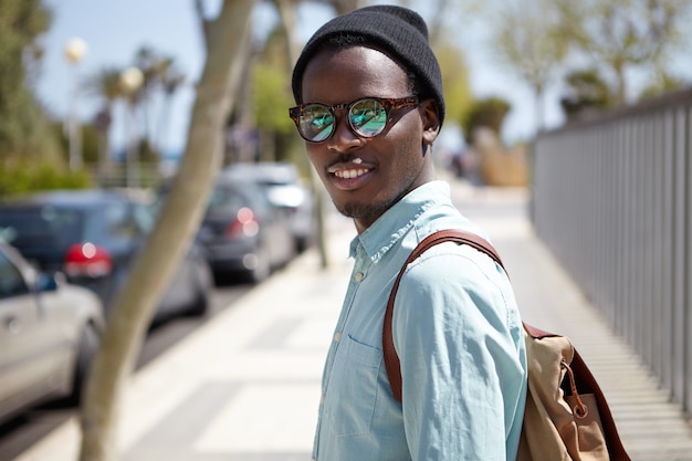 Urban summer portrait of handsome trendy looking young African American man in eyewear and hat walking on streets of foreign city, sightseeing, visiting landmarks while spending holidays abroad
