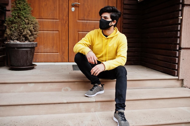 ファッショナブルな黄色のスウェットシャツを着た都会の若いヒップスターのインド人クールな南アジアの男は、新しい通常の間にパーカーと黒い顔の保護マスクを着用します