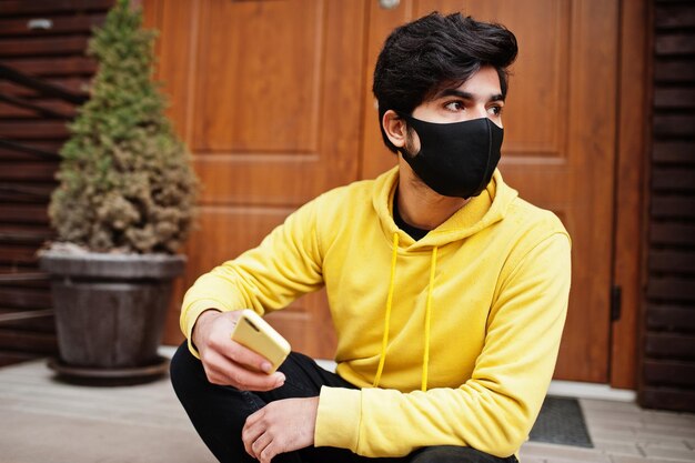 Urban young hipster indian man in a fashionable yellow sweatshirt with mobile phone Cool south asian guy wear hoodie and black face protect mask during new normal
