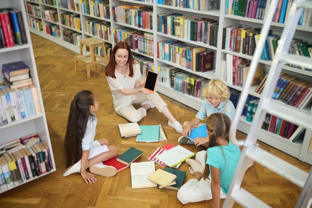 유용한 정보. 도서관에서 책과 공책을 들고 바닥에 앉아 있는 학교 아이들에게 태블릿을 보여주는 웃고 있는 젊은 빨간 머리 여성