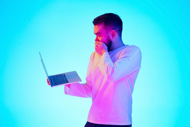 Free photo using laptop. caucasian man's portrait with device on blue studio background in neon light. beautiful male model. concept of human emotions, facial expression, sales, ad. copyspace for ad. close up