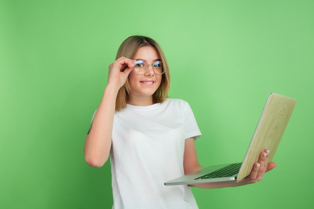 Using laptop emotional. Caucasian young woman's portrait isolated on green wall. Beautiful female model in white shirt.