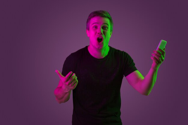 Using smartphone, betting, winning. Caucasian man's portrait on purple studio background in neon light. Beautiful male model in black shirt. Concept of human emotions, facial expression, sales, ad.