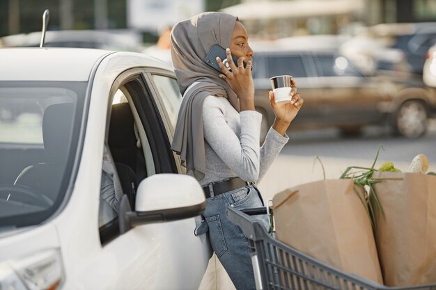 待っている間にスマートフォンを使用します。昼間の電気自動車充電ステーションのアフリカ民族の女性。真新しい車。