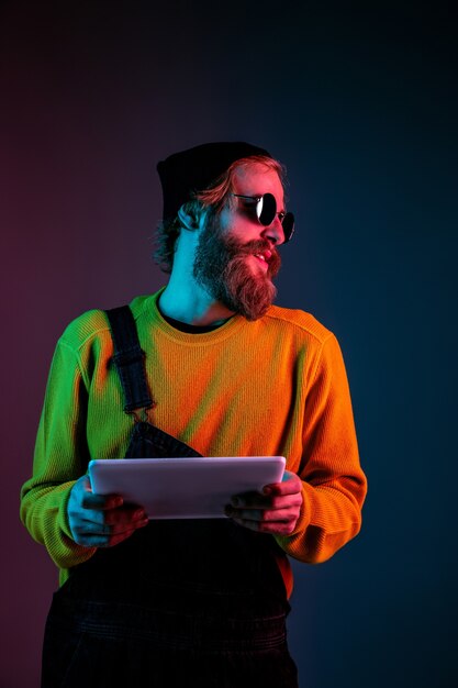 Using tablet, looks happy. Caucasian man's portrait on gradient studio background in neon light. Beautiful male model with hipster style. Concept of human emotions, facial expression, sales, ad.