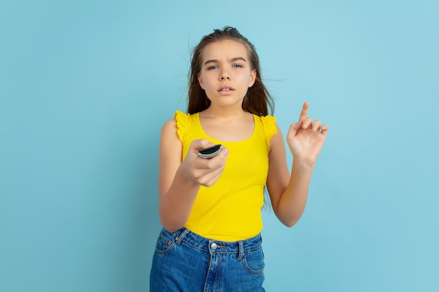 Using TV remote. Caucasian teen girl's portrait isolated on blue