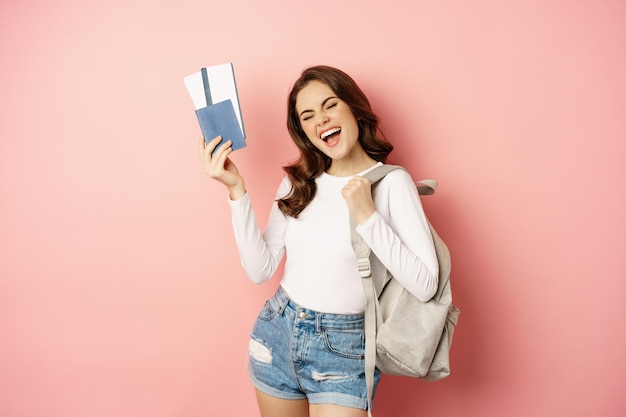Vacation and holiday. Beautiful girl going on trip, holding passport with airplane tickets, holding backpack, travelling, standing over pink background