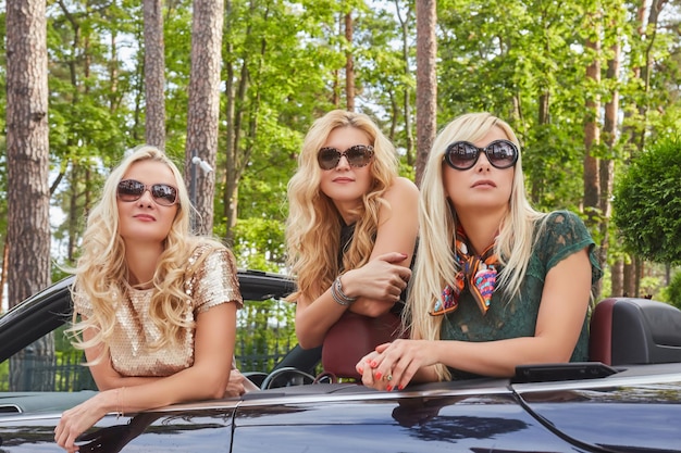 휴가와 사람들 개념입니다. 여름 여행 휴가에 카브리올레 차에 앉아 선글라스에 유행 옷을 입고 세 행복 한 금발 여자 친구.