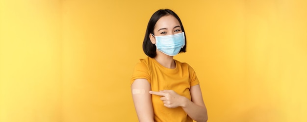 Vaccination and covid pandemic concept smiling asian woman in medical face mask showing her shoulder