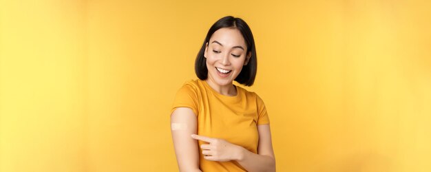 Концепция вакцинации и пандемии covid19 Счастливая и здоровая азиатка, указывающая на плечо пластырем после вакцинации от коронавируса желтого фона