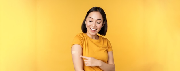 예방 접종 및 covid19 전염병 개념 코로나바이러스 노란색 배경에서 예방 접종을 한 후 반창고로 어깨를 가리키는 행복하고 건강한 아시아 소녀