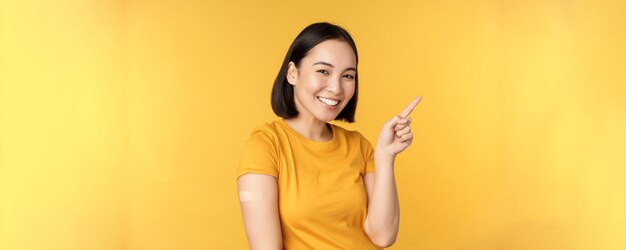 Vaccination and covid19 pandemic concept Smiling korean woman with band aid on shoulder after coronavirus vaccine shot pointing at banner with vaccinating campaign