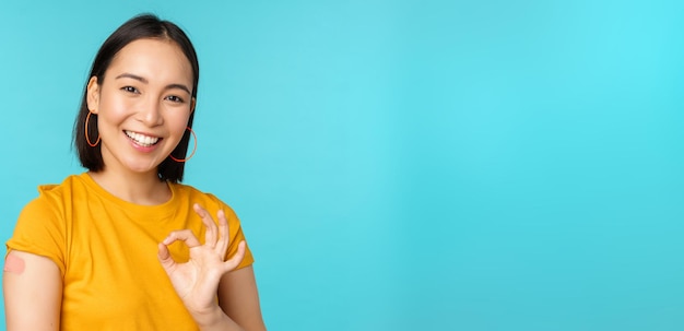 Free photo vaccine campaign from covid19 happy healthy asian girl showing okay sign and smiling bandaid on shoulder from coronavirus vaccination blue background