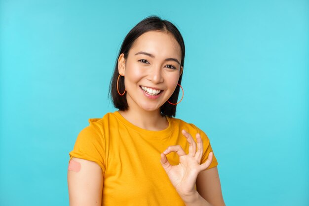 covid19의 백신 캠페인 코로나바이러스 백신 파란색 배경에서 어깨에 반창고를 하고 괜찮은 표시를 보여주는 행복한 건강한 아시아 소녀